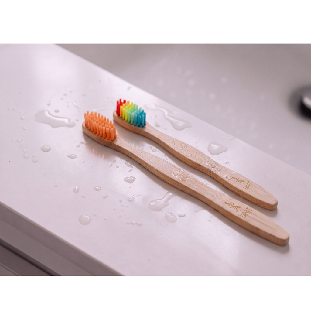 Two TANIT bamboo toothbrushes with rainbow bristles resting on a wet sink